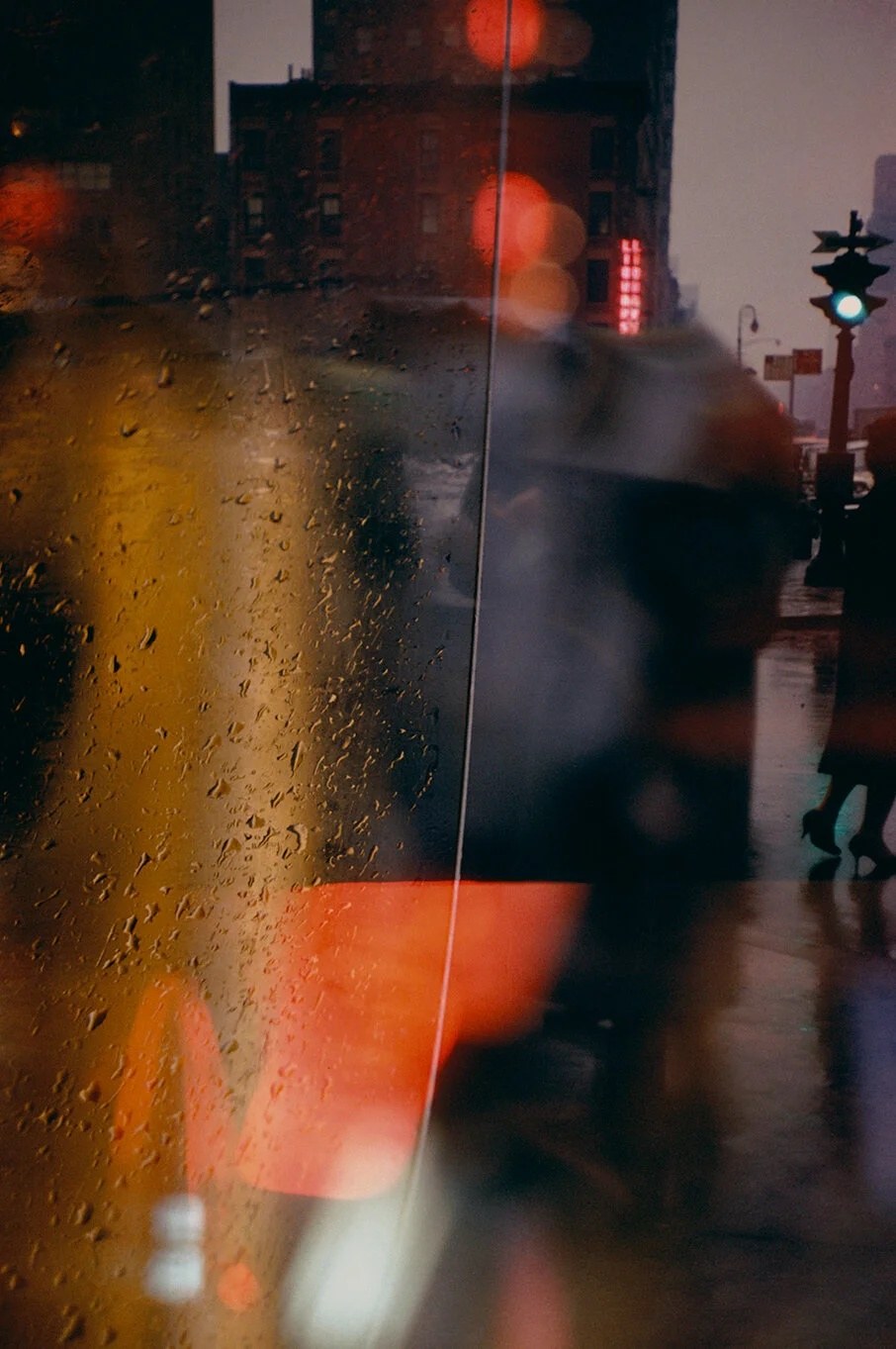 Rain-beaded glass with amber and red reflections, a green traffic light, and a woman&rsquo;s heels crossing at the far right.